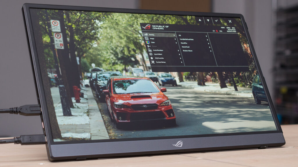 The 6 Best Portable Monitors for Content Creators in 2025 4 A low-angle shot of the 15.6-inch ASUS ROG Strix XG16AHP portable gaming monitor. The monitor is displaying an image of a red car parked on a tree-lined street. The on-screen display (OSD) menu is visible on the right side of the screen, with the "Gaming" option highlighted. The stand is visible in the background and a black cable is plugged into the left side of the monitor. The ROG logo is on the bottom bezel.