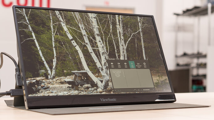 The 6 Best Portable Monitors for Content Creators in 2025 6 A slightly low-angle shot of the 15.6-inch ViewSonic VG1655 portable monitor. The monitor is sitting on a desk with its integrated kickstand unfolded, and it has a soft black case attached. The screen displays a nature photo of a picnic table with a forest and white birch trees in the background. The monitor's on-screen display (OSD) is visible on the right side of the screen, with the "ViewMode" menu highlighted.