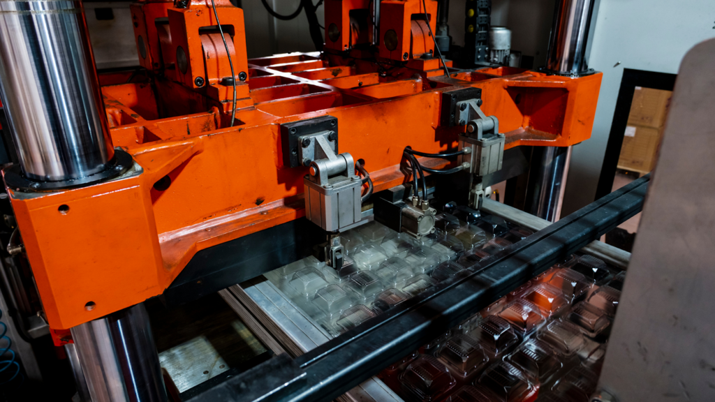 Smart, Sustainable, and Local: Why Modern Thermoforming Is Redefining Packaging Strategy 1 A large orange industrial thermoforming machine forming clear plastic food containers on a conveyor belt in a manufacturing facility.