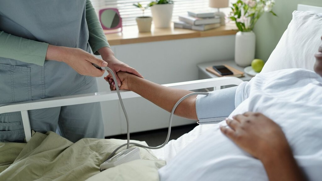 A healthcare professional in green scrubs checking the blood pressure of a patient in a hospital bed using a digital monitor, highlighting modern internal medicine.
