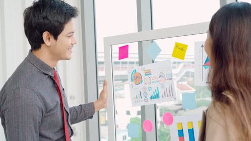 A group of marketing or UX professionals reviews concepts on a glass whiteboard covered in sticky notes and data charts. A person on the right, wearing a light blue shirt and headphones around their neck, thoughtfully rests their chin on their hand while a colleague points to a mobile app wireframe on the board.