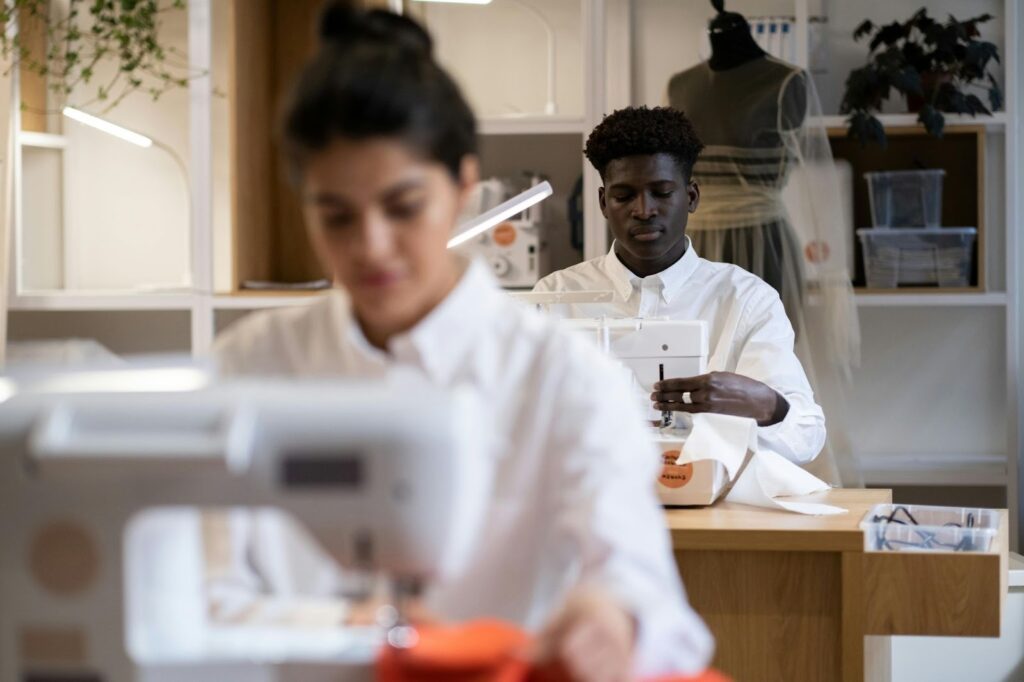 A focused shot of two fashion professionals in white shirts working diligently at industrial sewing machines in a brightly lit apparel manufacturing facility.