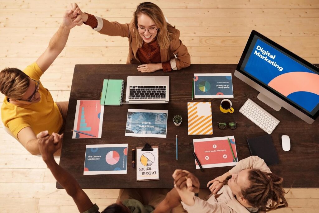 How to Scale Paid Campaigns Without Burning Budget 1 A diverse marketing team celebrating around a wooden conference table filled with performance reports and social media analytics. A computer monitor displays "Digital Marketing," while the team members join hands in a gesture of unity and success.