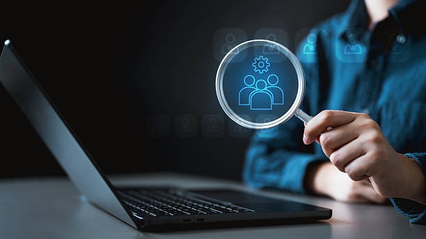 A close-up of a person using a magnifying glass over a laptop screen to inspect a glowing blue icon of people with a gear, representing technical SEO, user data analysis, and audience targeting.