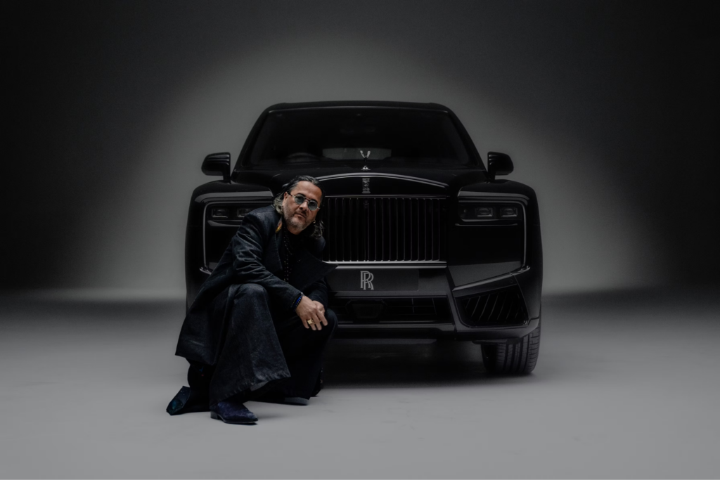 Elevated Automotive: The Pinnacle of Motoring Excellence 1 A man with long dark hair, wearing a long black coat and round sunglasses, crouches in front of a black Rolls-Royce Cullinan. The SUV's iconic chrome grille with the 'RR' badge is prominent behind him in a darkly lit, minimalist studio setting against a gray backdrop.