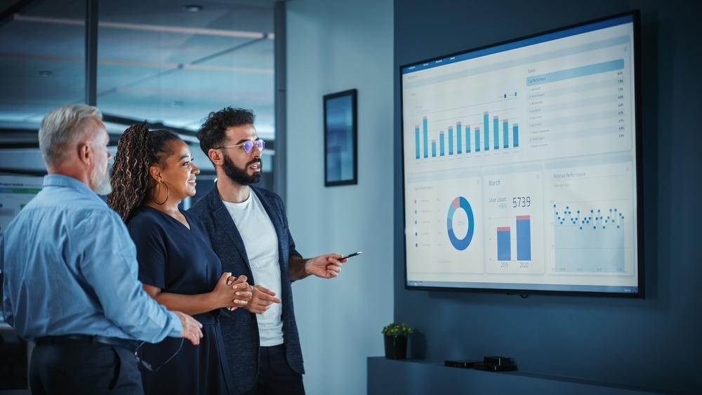 How Digital Marketing Agencies Help UAE Businesses Generate Consistent Leads in Competitive Markets 1 A group of diverse business professionals analyzing performance metrics, charts, and bar graphs displayed on a large digital monitor in a modern office meeting room