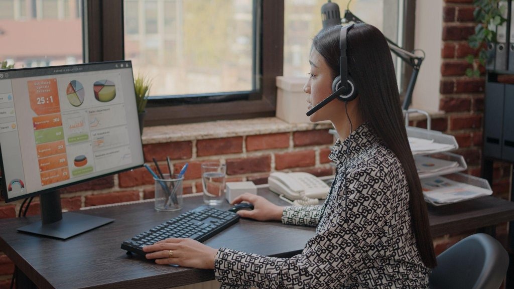 How Virtual Assistant Task Services Help Small Businesses Improve Productivity 1 A virtual assistant wearing a headset and working at a desk with a computer displaying analytics charts and performance metrics.